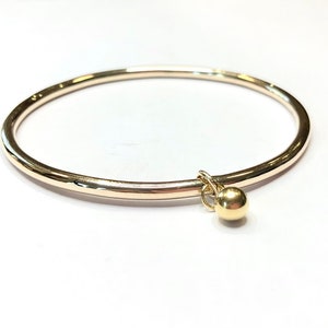 May include: A gold bangle bracelet with a small gold ball charm hanging from it.