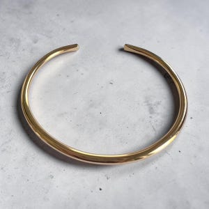 May include: A polished gold-tone cuff bracelet with a smooth, rounded surface and a wide opening at the top. The metal band features a consistent thickness throughout its circular shape, reflecting light across its curved, reflective surface.