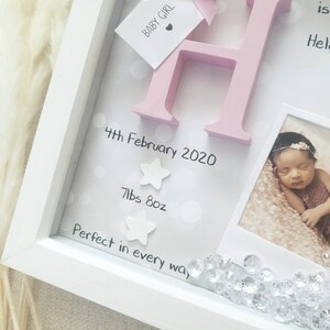 Op de afbeelding: Een witte schaduwdooslijst met een roze houten letter H, een label met BABY GIRL en een foto van een pasgeborene. De achtergrond bevat witte stippen, de tekst 4th February 2020, 7lbs 8oz, Perfect in every way en twee witte sterren.