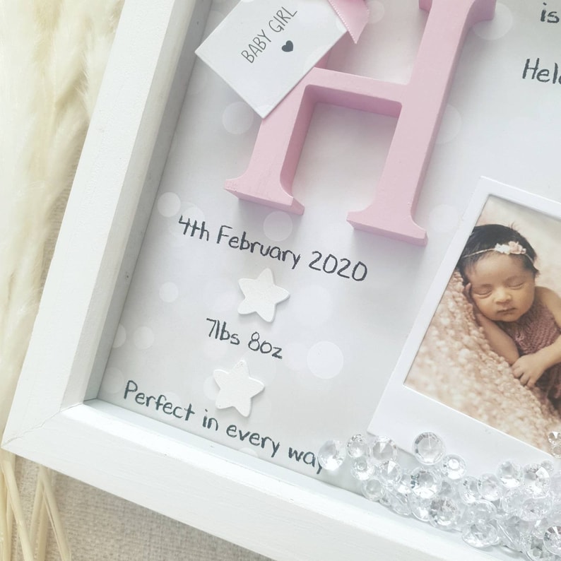 Op de afbeelding: Een witte schaduwdooslijst met een roze houten letter H, een label met BABY GIRL en een foto van een pasgeborene. De achtergrond bevat witte stippen, de tekst 4th February 2020, 7lbs 8oz, Perfect in every way en twee witte sterren.