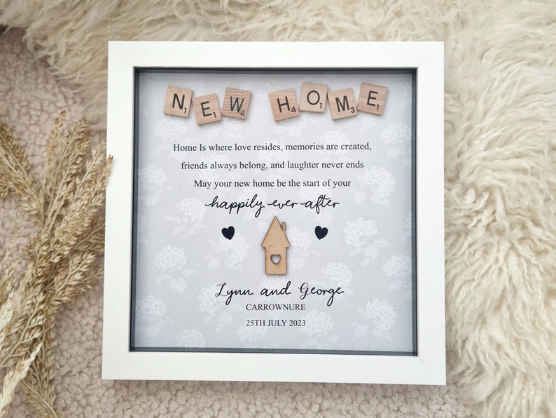 May include: A white framed picture with a gray background and a white floral pattern. The words "NEW HOME" are spelled out with wooden letter tiles. The text reads "Home is where love resides, memories are created, friends always belong, and laughter never ends. May your new home be the start of your happily ever after." The names "Lynn and George" are written in cursive, along with the date "25TH JULY 2023".