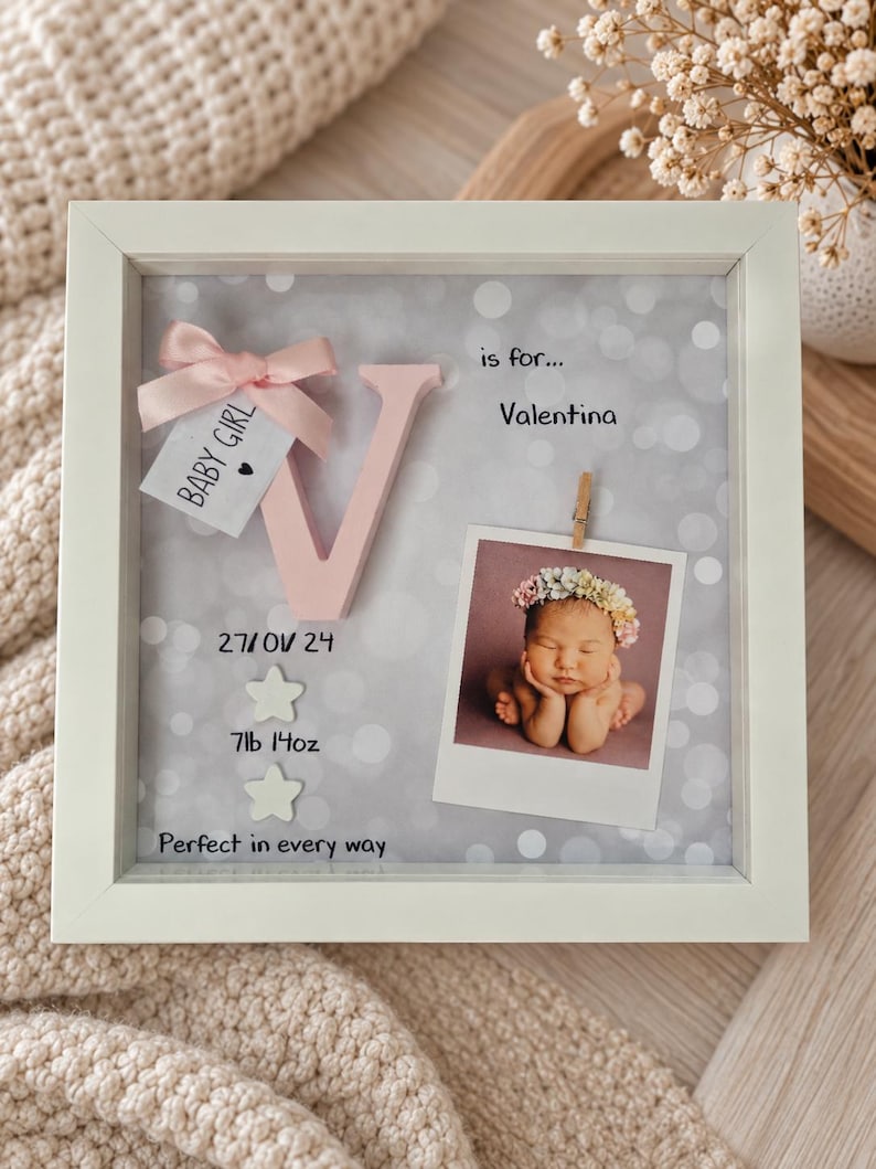 Op de afbeelding: Een witte vierkante schaduwdooslijst met een roze letter V, een kleine roze strik en een label met BABY GIRL. Het bevat een babyfoto, de tekst is for... Valentina, 27/01/24, 7lb 14oz, en Perfect in every way, met twee ster-pictogrammen.