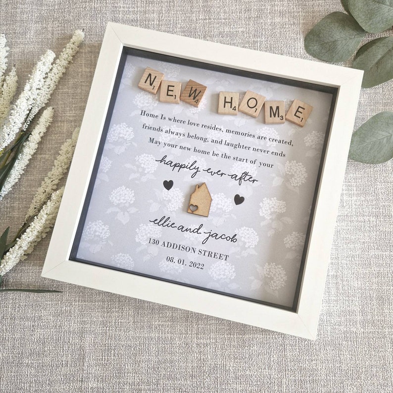 May include: A white picture frame with a gray background and a floral pattern. The frame contains the words "NEW HOME" spelled out with wooden letter tiles. The text reads "Home is where love resides, memories are created, friends always belong, and laughter never ends. May your new home be the start of your happily ever after." The names "ellie and jacob" are written below the text, along with the address "130 ADDISON STREET" and the date "08.01.2022".
