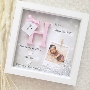 Op de afbeelding: Een witte schaduwdooslijst met een grote roze letter H, een klein label met BABY GIRL en een roze strik, een foto van een pasgeborene, twee witte sterren en doorzichtige kristallen. Tekst: is for... Helena Elizabeth, 4th February 2020, 7lbs 8oz, Perfect in every way.