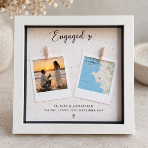 May include: A white square shadow box frame containing a light-coloured background with small heart patterns. It displays two polaroid-style photos clipped to the backing, one showing a beach proposal and the other a map of Paphos, Cyprus. Text reads Engaged, Olivia & Jonathan, and Paphos, Cyprus, 12th September 2026.