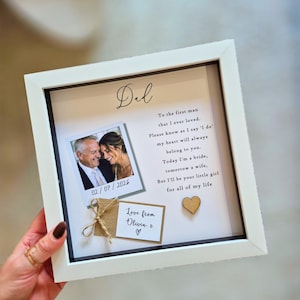 May include: A white square shadow box frame containing a printed message addressed to Dad, a polaroid-style photo of a man and a woman, a small kraft paper tag tied with twine reading Love from Olivia x, and a small wooden heart embellishment.