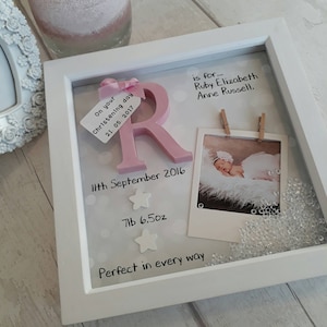 Op de afbeelding: Een witte schaduwdooslijst met een roze houten letter R, een babyfoto bevestigd met houten knijpers, twee witte sterren en tekst met de naam Ruby Elizabeth Anne Russell, de datum 11 september 2016 en het gewicht 7lb 6.5oz.