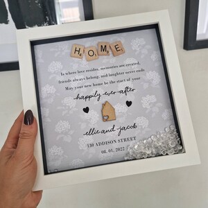 May include: A white picture frame with a gray background and a floral pattern. The frame contains a message about home, love, and memories. The message is written in black lettering and includes the words "HOME", "happily ever after", and "ellie and jacob". The frame also includes a small wooden house cutout and a scattering of clear rhinestones.