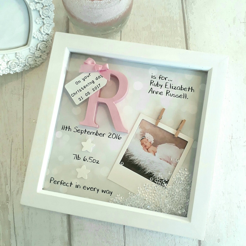 Op de afbeelding: Een vierkante witte schaduwdooslijst met een roze letter R, een klein label met een roze strik, een babyfoto, twee kleine witte sterren en losse doorzichtige kristallen. De tekst luidt: On your Christening day 21. 05. 2017, is for... Ruby Elizabeth Anne Russell, 11th September 2016, 7lb 6.5oz, Perfect in every way.