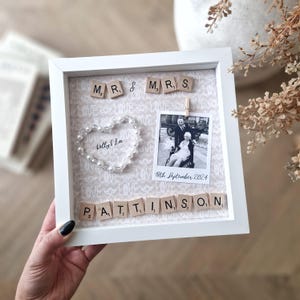 May include: White frame with a light brown background featuring a black and white photo of a couple, a pearl heart, and the words "MR & MRS" and "PATTINSON" spelled out with wooden letter tiles. The date "8th September 2024" is written below the photo.