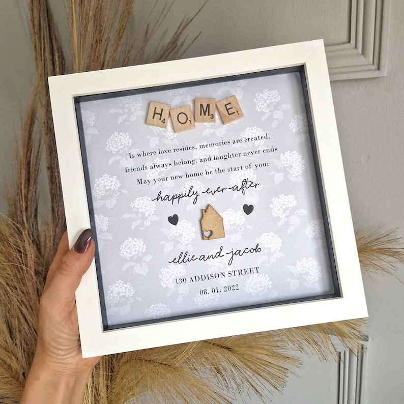May include: A white framed picture with a gray background and a white floral pattern. The picture has the word "HOME" spelled out with wooden letter tiles. The text reads "Is where love resides, memories are created, friends always belong, and laughter never ends. May your new home be the start of your happily ever after." The names "ellie and jacob" are written below the text, along with the address "130 ADDISON STREET" and the date "08.01.2022".