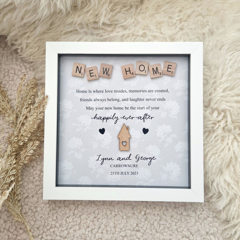 May include: A white picture frame with a gray background and a message that reads "NEW HOME" in wooden letter tiles. The message continues with "Home is where love resides, memories are created, friends always belong, and laughter never ends. May your new home be the start of your happily ever after." The names "Lynn and George" are written below the message, along with the location "CARROWNURE" and the date "25TH JULY 2023".