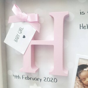 Op de afbeelding: Een lichtroze houten letter H in een schaduwdooslijst. Een klein wit label met de tekst BABY GIRL en een hartpictogram is met een roze satijnen lint aan de letter bevestigd. De tekst 4th February 2020 staat onder de letter gedrukt.