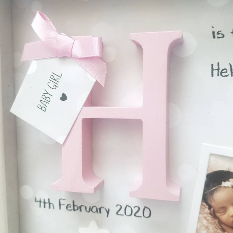 Op de afbeelding: Een lichtroze houten letter H in een schaduwdooslijst. Een klein wit label met de tekst BABY GIRL en een hartpictogram is met een roze satijnen lint aan de letter bevestigd. De tekst 4th February 2020 staat onder de letter gedrukt.