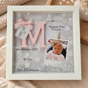 Op de afbeelding: Een witte schaduwdooslijst met een grote roze letter M, een babyfoto en tekst waaronder "is for... Margaux Mary Priscott", "6th February 2024", "7lb 15oz" en "Perfect in every way". Een klein labeltje toont "BABY GIRL" met een roze strik.