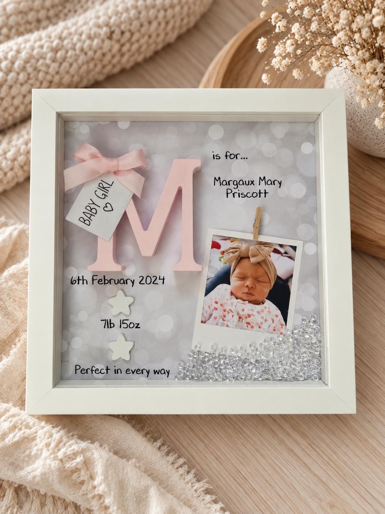 Op de afbeelding: Een witte schaduwdooslijst met een grote roze letter M, een babyfoto en tekst waaronder "is for... Margaux Mary Priscott", "6th February 2024", "7lb 15oz" en "Perfect in every way". Een klein labeltje toont "BABY GIRL" met een roze strik.