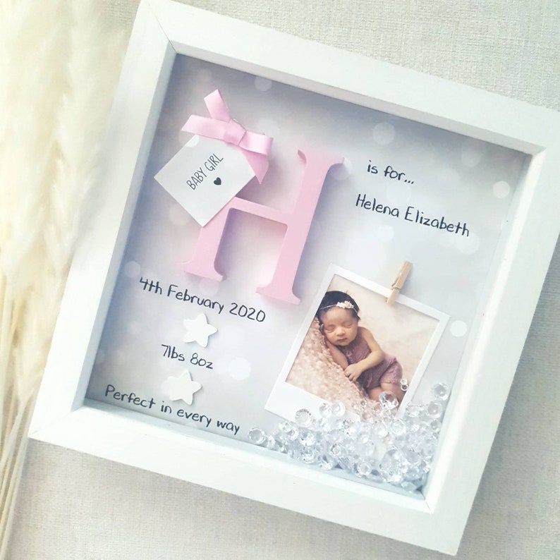 Op de afbeelding: Een witte schaduwdooslijst met een grote roze letter H, een label met een roze strik met de tekst BABY GIRL, een foto van een pasgeborene, twee witte sterren en doorzichtige kristallen. De tekst bevat 4th February 2020, 7lbs 8oz, is for... Helena Elizabeth en Perfect in every way.