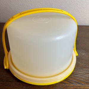 May include: A round, off-white cake carrier with a bright yellow handle and base. The carrier is made of translucent plastic, with a yellow handle that arches over the top. The base is also yellow, with a cream-colored ring around the edge.