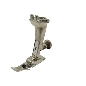 May include: A silver sewing machine foot with a number "2" on the side. The foot has a curved needle and a small screw on the side.