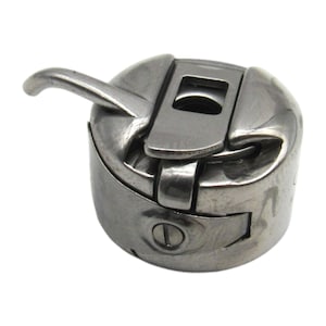 May include: A silver sewing machine bobbin case with a lever on the side. The bobbin case is designed to hold a bobbin of thread for sewing.