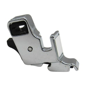 May include: A silver sewing machine foot with a black plastic piece on the side. The foot has a small hole in the center and a small metal plate on the side.