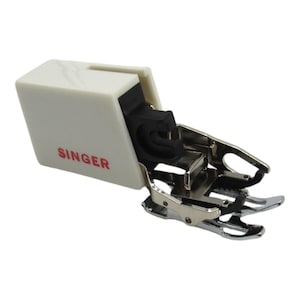 May include: A sewing machine presser foot with a white plastic housing and the red word "SINGER" printed on it. The foot has a black rubber component and a shiny, silver-colored metal base with two prongs.
