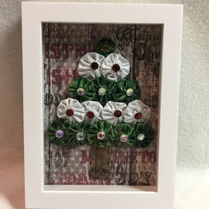 May include: A framed Christmas tree made of white and green fabric rosettes with red and iridescent rhinestones. The background is a vintage-style paper with the words "Best Christmas Ever" and "Merry & Bright".