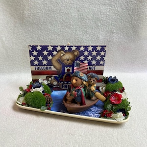 May include: A decorative figurine depicting three teddy bears in a boat, set against a backdrop of the American flag with the words "FREEDOM NOT". The bears are dressed in patriotic outfits, with floral arrangements.