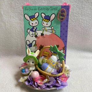 Op de afbeelding: Een paaswenskaart met een decoratief arrangement. De kaart toont cartoonkonijntjes en de tekst "for You at Easter Time". Een mand met versierde eieren en een konijnenfiguur ligt op een bed van paarse en roze bloemen.