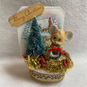 Pode incluir: Decoração de Natal vintage com um rato de cerâmica com um laço vermelho, sentado numa base cheia de fios dourados. Inclui uma pequena árvore de Natal verde e um cenário com uma cena de inverno. O letreiro diz "Merry Christmas".