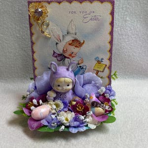 May include: An Easter-themed decorative piece featuring a vintage card with a child in a bunny costume. The card is adorned with a gold flower pin and set on a bed of purple and white flowers, with a small figurine in a bunny outfit.