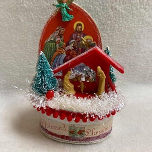 May include: A vintage Christmas decoration featuring a nativity scene inside a red-roofed structure. The base is decorated with red beads and the words "Blessed Christmas". A green angel figurine is perched above the scene.