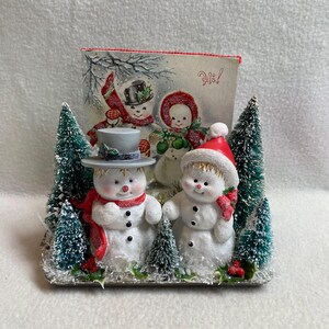 Könnte beinhalten: Eine weihnachtliche Figur mit zwei Schneemännern mit roten Schals und Hüten und einer Vintage-Karte mit Schneemännern und dem Wort "Hi!". Die Schneemänner sind von kleinen Tannenbäumen umgeben.