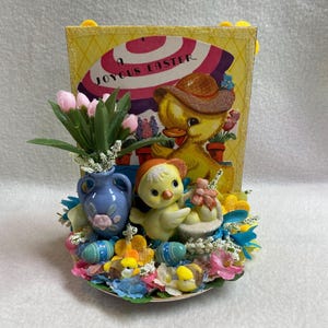 May include: A vintage Easter decoration featuring a yellow card with a duck illustration and the text "A Joyous Easter." The arrangement includes a blue vase with pink tulips, ceramic chicks, and colorful floral accents, with painted Easter eggs.