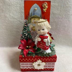 Puede incluir: Un adorno navideño que presenta una figura de ángel de porcelana con un halo dorado que sostiene un calcetín rojo con la inscripción Merry Christmas. La base es una caja roja con borde de encaje y una flor blanca, decorada con un árbol y flores.