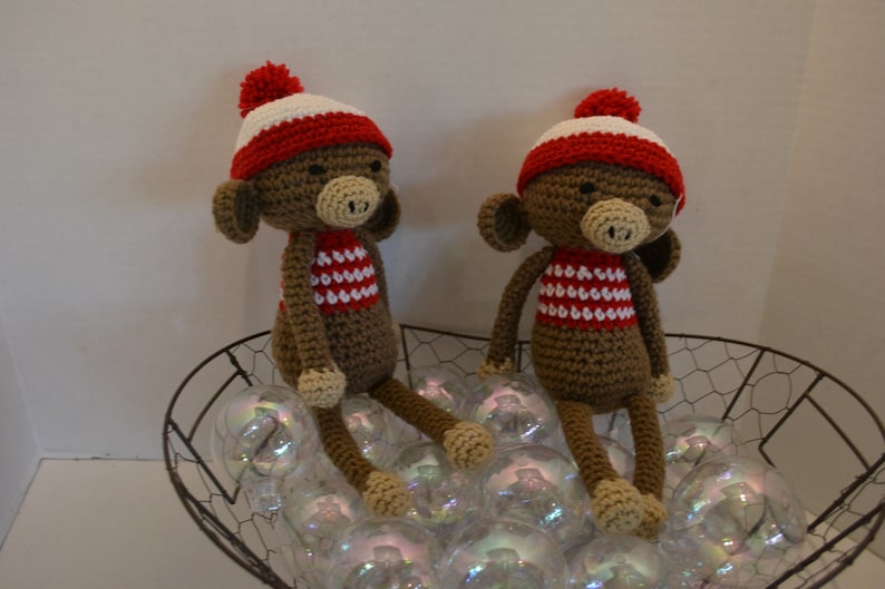 May include: Two brown crocheted monkeys wearing red and white striped shirts and red and white knit hats with white pom-poms. The monkeys are sitting in a wire basket filled with iridescent glass ornaments.