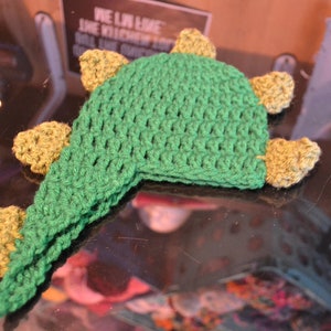 Puede incluir: Un gorro de dinosaurio de ganchillo verde hecho a mano con detalles amarillos. El gorro presenta una cabeza puntiaguda y peque&ntilde;as p&uacute;as triangulares a lo largo de la espalda. El gorro est&aacute; dise&ntilde;ado para la cabeza de un ni&ntilde;o y est&aacute; hecho de hilo suave.