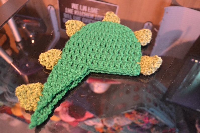 Puede incluir: Un gorro de dinosaurio de ganchillo verde hecho a mano con detalles amarillos. El gorro presenta una cabeza puntiaguda y peque&ntilde;as p&uacute;as triangulares a lo largo de la espalda. El gorro est&aacute; dise&ntilde;ado para la cabeza de un ni&ntilde;o y est&aacute; hecho de hilo suave.