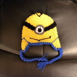 Puede incluir: Un gorro amarillo tejido a mano con detalles negros y un dise&ntilde;o de ojo gris y blanco. El gorro presenta orejeras azules y lazos trenzados con borlas. Este gorro est&aacute; inspirado en un popular personaje animado.