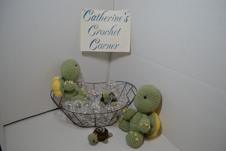 Pu&ograve; includere: Tre tartarughe all'uncinetto, due verdi e una marrone, in un cesto di filo metallico pieno di bolle di plastica trasparenti. Il cartello sopra dice "Catherine's Crochet Corner."
