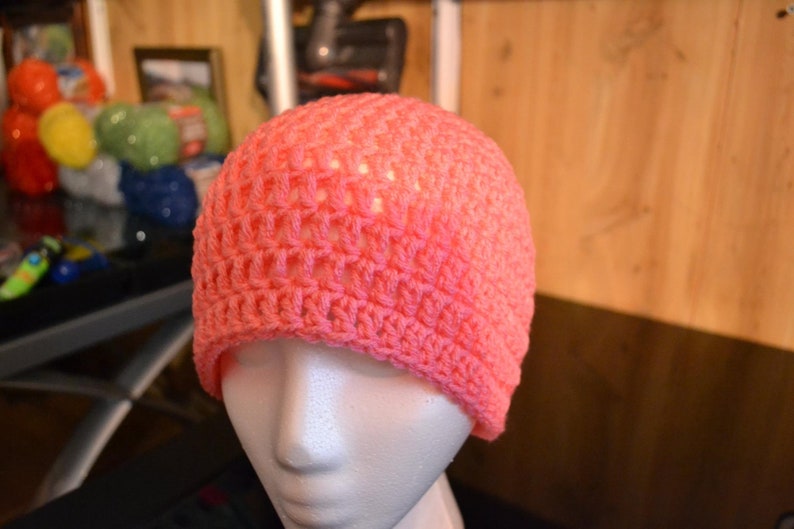 Puede incluir: Un gorro tejido a ganchillo de color coral. El gorro se exhibe en una cabeza de maniqu&iacute;, mostrando su dise&ntilde;o de tejido abierto. El color del gorro es un tono vibrante y c&aacute;lido, perfecto para a&ntilde;adir un toque de color a cualquier atuendo.