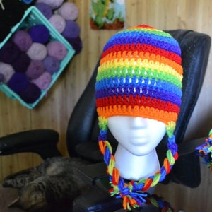 Puede incluir: Un gorro de ganchillo hecho a mano con un vibrante patr&oacute;n de arco&iacute;ris. El gorro tiene orejeras con lazos trenzados y una borla colorida. El gorro se exhibe en una cabeza de maniqu&iacute;. Hilo de varios colores es visible en el fondo.