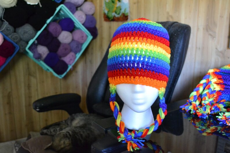 Puede incluir: Un gorro de ganchillo hecho a mano con un vibrante patr&oacute;n de arco&iacute;ris. El gorro tiene orejeras con lazos trenzados y una borla colorida. El gorro se exhibe en una cabeza de maniqu&iacute;. Hilo de varios colores es visible en el fondo.