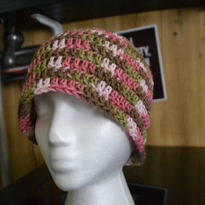 Puede incluir: Un gorro de ganchillo hecho a mano con un patr&oacute;n de rayas rosas, verdes y marrones. El gorro se muestra en una cabeza de maniqu&iacute; blanca. El gorro es de estilo gorro, adecuado para climas m&aacute;s fr&iacute;os. El hilo parece suave y las puntadas son apretadas.