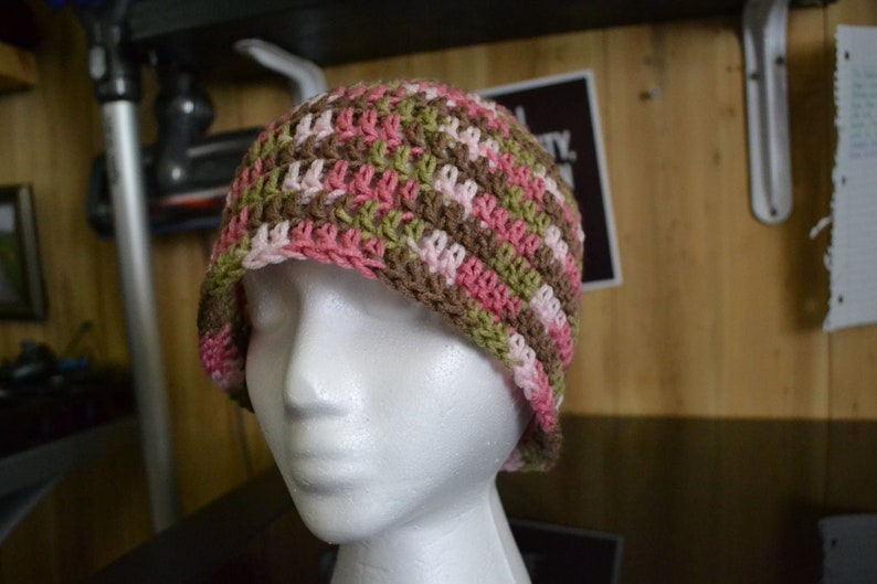 Puede incluir: Un gorro de ganchillo hecho a mano con un patr&oacute;n de rayas rosas, verdes y marrones. El gorro se muestra en una cabeza de maniqu&iacute; blanca. El gorro es de estilo gorro, adecuado para climas m&aacute;s fr&iacute;os. El hilo parece suave y las puntadas son apretadas.