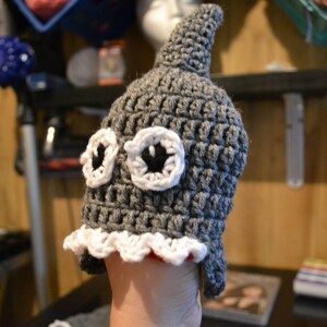Puede incluir: Un gorro de ganchillo gris hecho a mano con aleta de tibur&oacute;n y grandes ojos con bordes blancos y centros negros. El gorro tiene un ribete festoneado blanco y rojo. Un accesorio caprichoso.