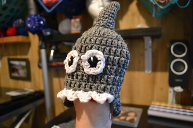 Puede incluir: Un gorro de ganchillo gris hecho a mano con aleta de tibur&oacute;n y grandes ojos con bordes blancos y centros negros. El gorro tiene un ribete festoneado blanco y rojo. Un accesorio caprichoso.