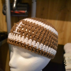 Puede incluir: Un gorro de ganchillo marr&oacute;n y blanco dise&ntilde;ado para parecerse a un bal&oacute;n de f&uacute;tbol americano. El gorro presenta hilo marr&oacute;n con rayas blancas y detalles de costura blancos, creando un aspecto deportivo. El gorro se muestra en una cabeza de maniqu&iacute; blanca.