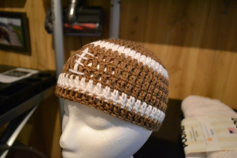 Puede incluir: Un gorro de ganchillo marr&oacute;n y blanco dise&ntilde;ado para parecerse a un bal&oacute;n de f&uacute;tbol americano. El gorro presenta hilo marr&oacute;n con rayas blancas y detalles de costura blancos, creando un aspecto deportivo. El gorro se muestra en una cabeza de maniqu&iacute; blanca.
