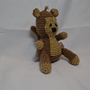 May include: A brown and tan crocheted monkey toy with black eyes. The monkey is sitting on a white surface.
